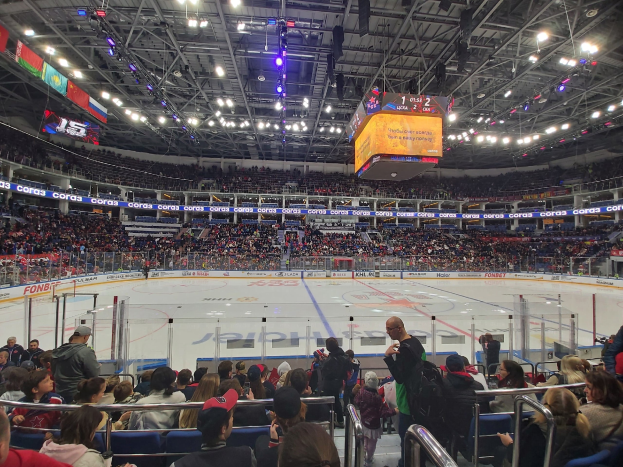 Eine große Menschenmenge in einer Hockeyarena, einige sitzen und einige stehen, schaut ein Spiel unter hellen Lichtern mit Bannern an der Decke und einem Zaun um die Eisfläche.