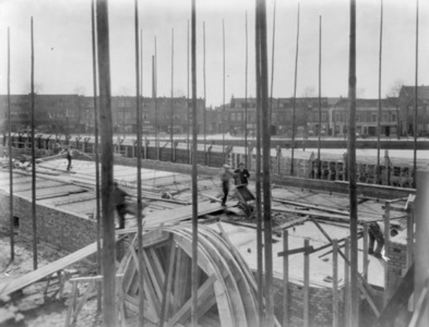 Schwarz-weiß-Foto einer Baustelle mit Arbeitern, hölzernen Pfählen, einem Rad im Vordergrund und Gebäuden, Bäumen und einem klaren Himmel im Hintergrund.