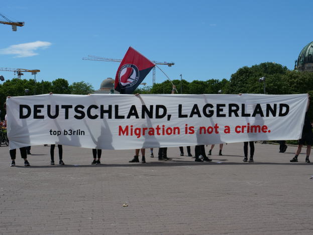 Eine Gruppe von Menschen mit einem Banner 'Deutschland, Lagerland Migration ist kein Verbrechen' und einer Flagge, die auf dem Boden stehen, mit Straßenlaternen, Bäumen, Kränen, einem Gebäude und einem bewölkten Himmel im Hintergrund.