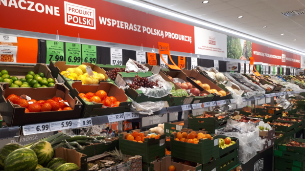 Ein Geschäft mit verschiedenen Obst- und Gemüsesorten in Kartons mit Preisschildern, Wandtafeln mit Text und Bildern sowie Deckenbeleuchtung.