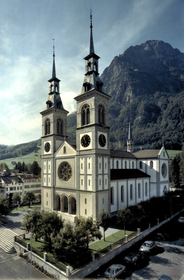 Alte Kirche mit zwei Türmen umgeben von Stadtgebäuden, Grünflächen, Fahrzeugen und Hügeln unter einem bewölkten Himmel.