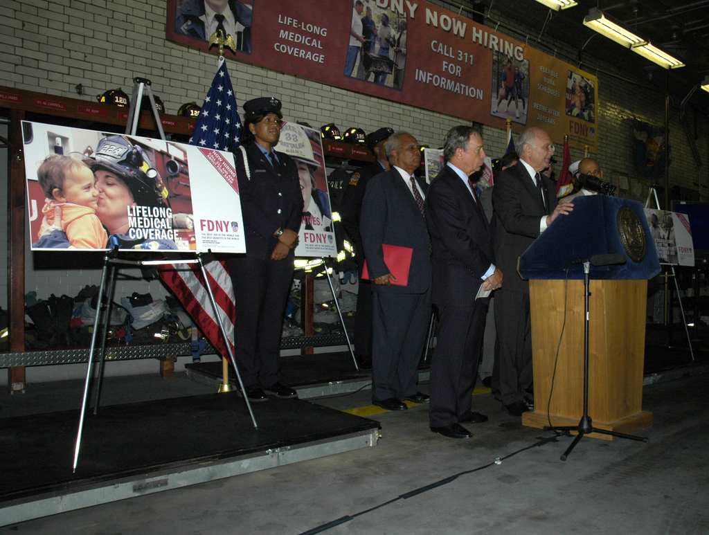 Eine Gruppe von Menschen steht in der Nähe eines Podiums mit Mikrofonen, umgeben von Schildern auf Ständern, Flaggen und Schildern an der Wand, alles unter hellen Lichtern.