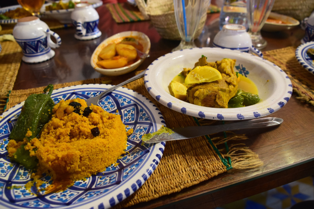 Ein Tisch mit Tellern voller Essen, Gläsern mit Wein, Schalen, Krügen, Besteck und Matten, mit Menschen im Hintergrund bei einem traditionellen marokkanischen Essen.