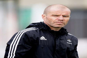 Bald coach in einer blauen Jacke mit weißen Streifen und weißem Kragen, der auf einem Fußballfeld steht und nachdenklich schaut.