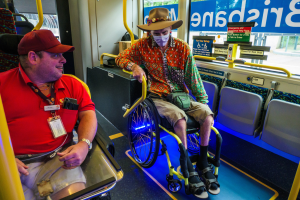 Zwei Männer im Rollstuhl in einem Bus, einer trägt ein rotes T-Shirt und eine rote Mütze, der andere ein orangefarbenes Hemd und einen braunen Hut, beide tragen Masken und Ausweise, mit gelben Stangen und Glasfenstern, durch die man Bäume, Gebäude und den Himmel sieht.