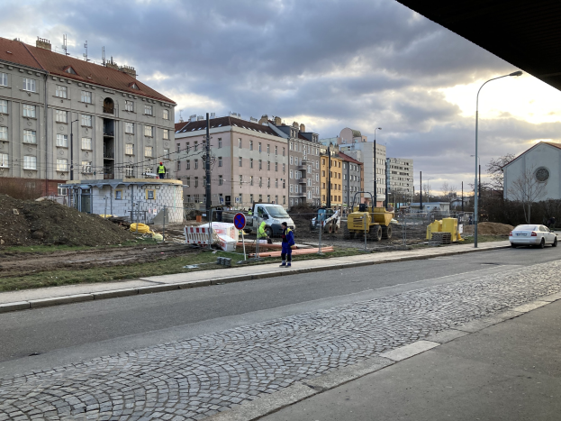 Eine Straße in der Stadt mit parkenden Autos, Gebäuden, Laternen, Bäumen und einem bewölkten Himmel, mit einer Sperre mit Fahrzeugen und Füßgängern im Vordergrund.