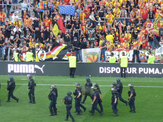 Eine Gruppe von Polizisten in Helmen und mit Waffen in der Hand läuft über ein Fußballfeld, im Hintergrund halten Menschen Fahnen und Transparente in der Nähe von Absperrungen.