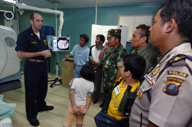 Eine Gruppe von Menschen in einem Krankenhauszimmer mit einem Mann, der ein Mikrofon hält, einem drapierten Bett, medizinischer Ausrüstung, einem Röntgengerät, einer Texttafel, einer Tür und Deckenleuchten.