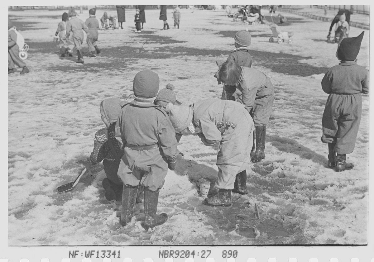 Eine Gruppe von Kindern mit Mützen und Schuhen spielt im Schnee, mit Menschen, Kinderwagen und anderen Gegenständen im schwarzen und weißen Hintergrund; Text ist unten im Bild sichtbar.