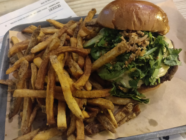 Ein Tablett mit einem Hamburger, der mit Salat, Tomate und Zwiebeln belegt und mit goldenen, knusprigen Pommes frites garniert ist, auf einem Tisch mit einem Blatt Papier daneben.