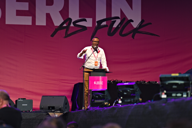 Ein Mann in einem weißen Hemd und braunen Hosen steht auf einer Bühne und hält ein Mikrofon in der Hand, mit einem pinken Banner im Hintergrund, auf dem "Berlin as Fuck" steht und Lautsprecher auf der Bühne.