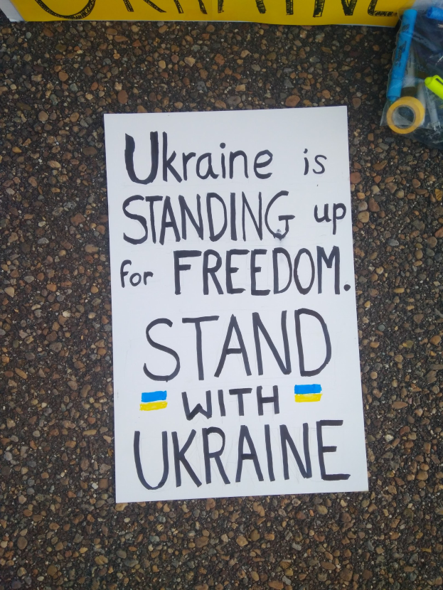 Weißer Zettel mit dem Text "Ukraine steht für Freiheit ein, steht mit Ukraine" auf dem Boden vor einer gelben Fahne, mit einer Plastikabdeckung mit verschiedenen Gegenständen rechts.