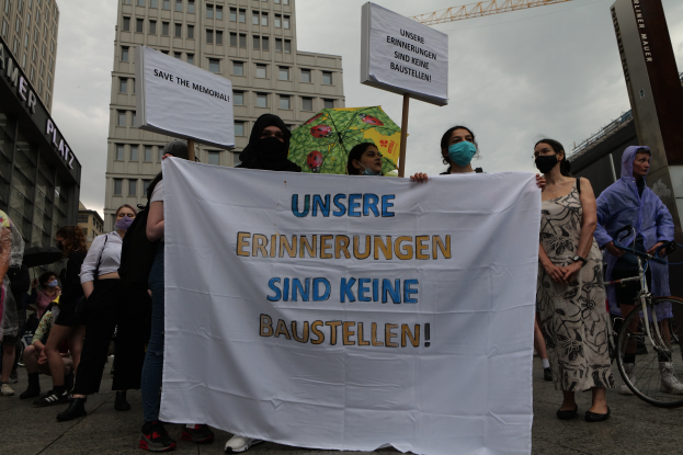 Eine Gruppe von Menschen mit Masken hält ein Banner mit der Aufschrift "Unsere Erinnerungen sind keine Baustellen" vor einem Gebäude, während einige ein Fahrrad, einen Schirm und Tafeln mit Text halten, und im Hintergrund Gebäude, ein Kran und ein bewölkter Himmel zu sehen sind.