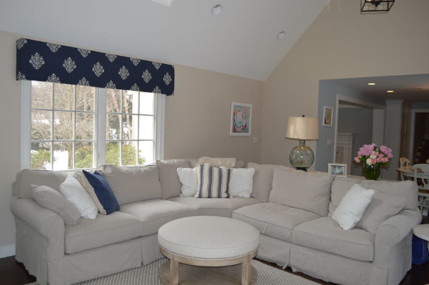 Ein Wohnzimmer mit einer Couch, Kissen, einem Tisch, Stühlen, einem Blumenstrauß, einem Foto Rahmen, einer Alarm Uhr und einem Fenster auf der linken Seite mit Deckenleuchten.