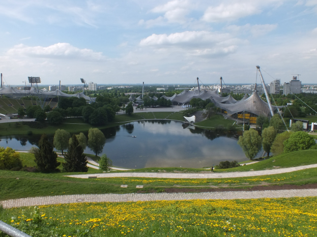 Blick auf das Olympiastadion von einem Hügel aus, mit einem Teich im Vordergrund, umgeben von Grün und gelben Blumen, und im Hintergrund sichtbare Gebäude unter einem klaren blauen Himmel.