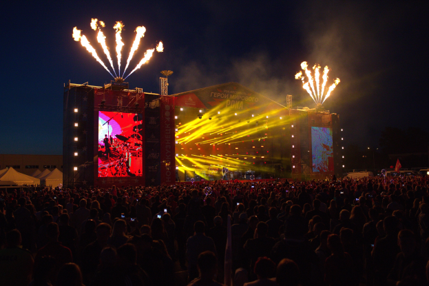 Eine große Menschenmenge versammelte sich auf einem Musikfestival, mit einer Bühne in der Mitte, auf der Musiker Instrumente spielen, beleuchtet von hellen Lichtern und umgeben von Bannern, Zelten links und einem dunklen Himmel mit Feuerwerk im Hintergrund.