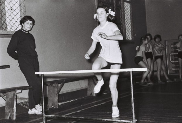 Schwarzes und weißes Foto einer Frau, die über ein Hindernis in einer Halle springt, mit Zuschauern und Bänken im Hintergrund.