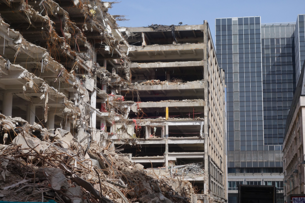 Ein großes Gebäude wird in einer von anderen Gebäuden, Fahrzeugen, Straßenelementen, Bäumen und einem klaren Himmel umgebenen Stadt abgerissen.