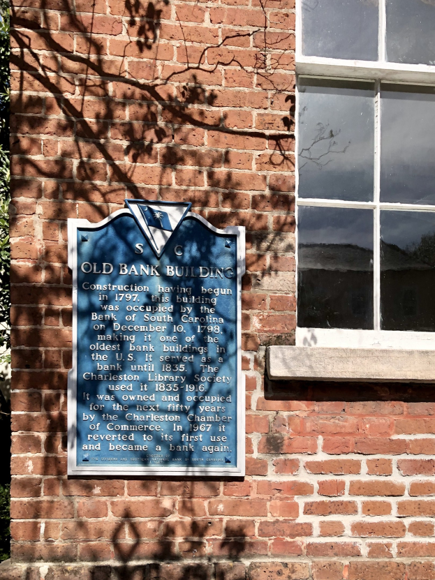 Altes Bankgebäude mit blauer Tafel an der Seite, Fenster rechts und Baum links.