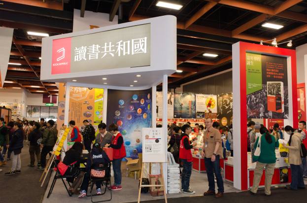 Eine große Gruppe von Menschen auf einer Messe vor einem Stand mit Stühlen, Texttafeln mit der Aufschrift "Book Republic Hong Kong Book Fair", Banner mit Bildern und Text sowie einer Decke mit Deckenleuchten.
