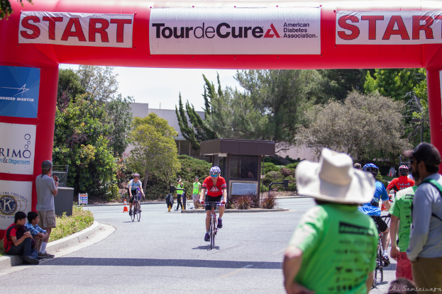 Eine belebte Straßenszene mit Fahrradfahrern, einigen auf einem Gehweg sitzend und anderen auf der Straße stehend, mit einer Hütte und umliegenden Pflanzen und Bäumen im Hintergrund.