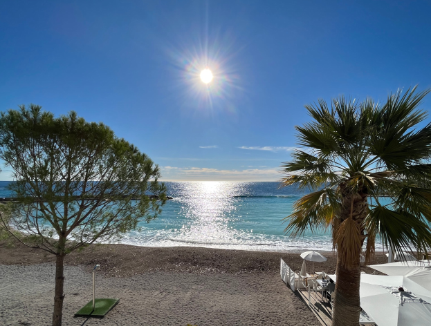 Ein sonniger Strand mit Palmen, Strandliegen und üppiger Vegetation, vor einem strahlend blauen Himmel mit der Sonne im Hintergrund.