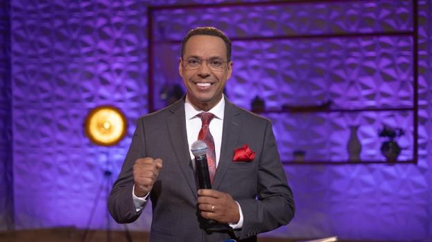 Stephen Colbert, Moderator von 'The Daily Show with Trevor Noah', steht auf einer Bühne und hält ein Mikrofon in der Hand, trägt einen Anzug, Krawatte und eine Brille, hinter ihm ist ein Licht und ein Ständer zu sehen.