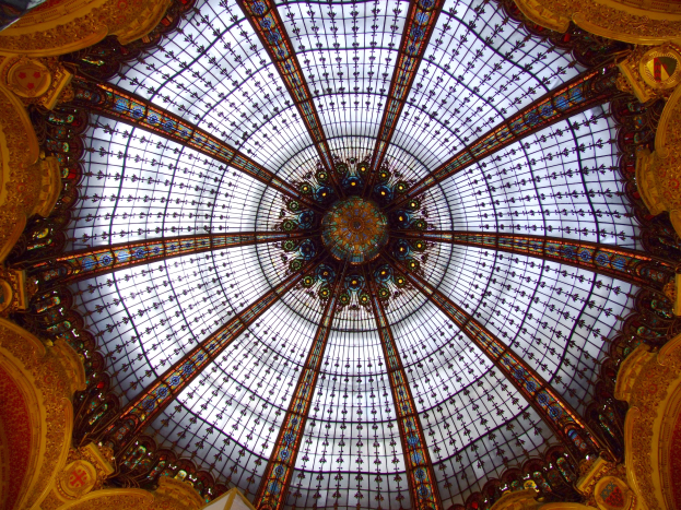 Ein Gebäudeinneres mit einer zentralen Glaskuppel mit detaillierten Verzierungen, umgeben von Wänden und Säulen, die das Licht in den Raum fallen lassen.