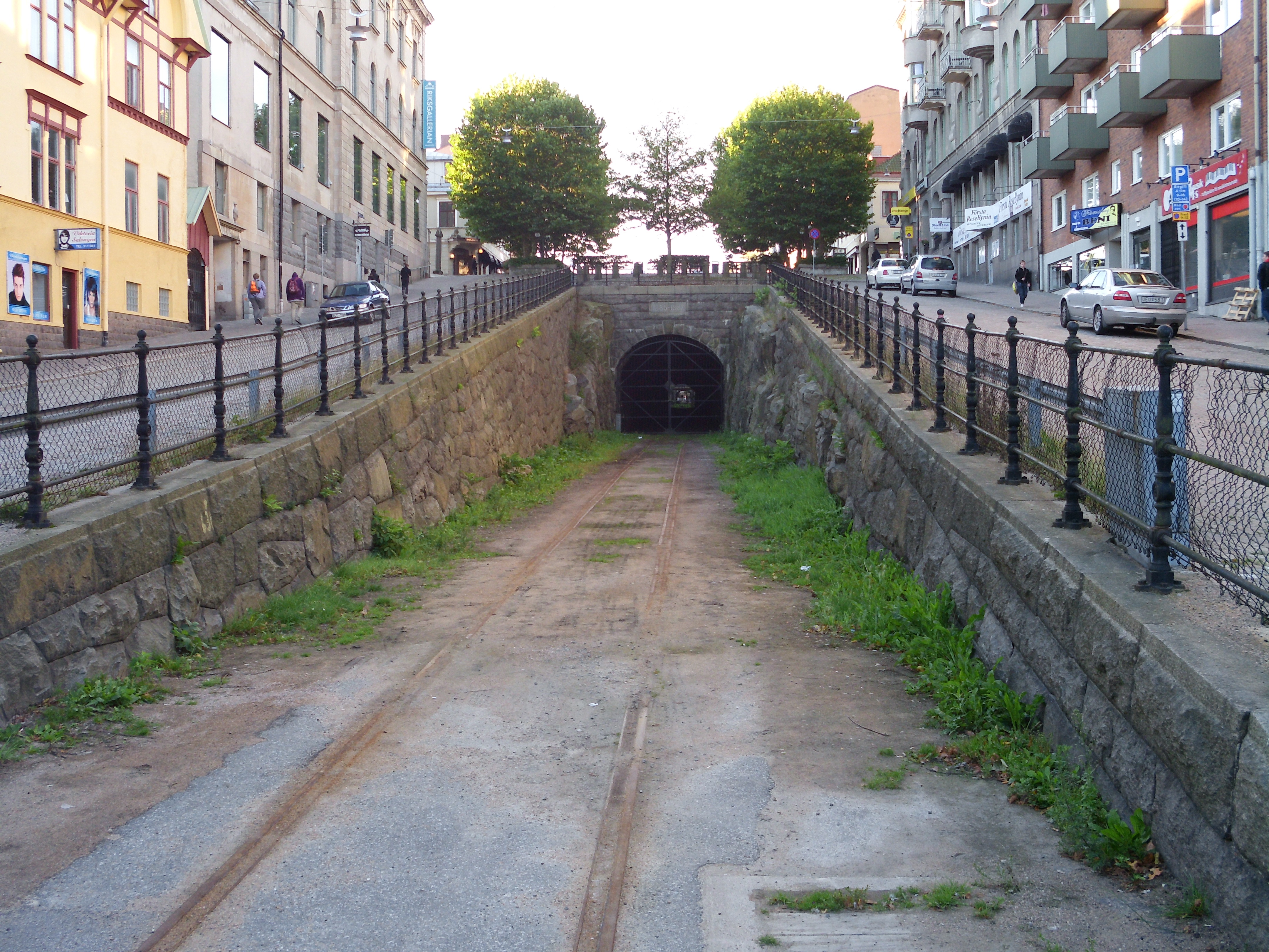 Ein kleiner Tunnel inmitten einer Stadtstraße, umgeben von Gras, einem Zaun, Fahrzeugen, Fußgängern, Gebäuden mit Fenstern, Bäumen, Schildern und einem klaren blauen Himmel.