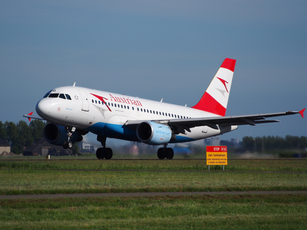 Airbus A320-200 der Austrian Airlines startet vom Frankfurter Flughafen, mit einem Schild im Vordergrund, Gras, Bäumen, Gebäuden und einem klaren blauen Himmel im Hintergrund.