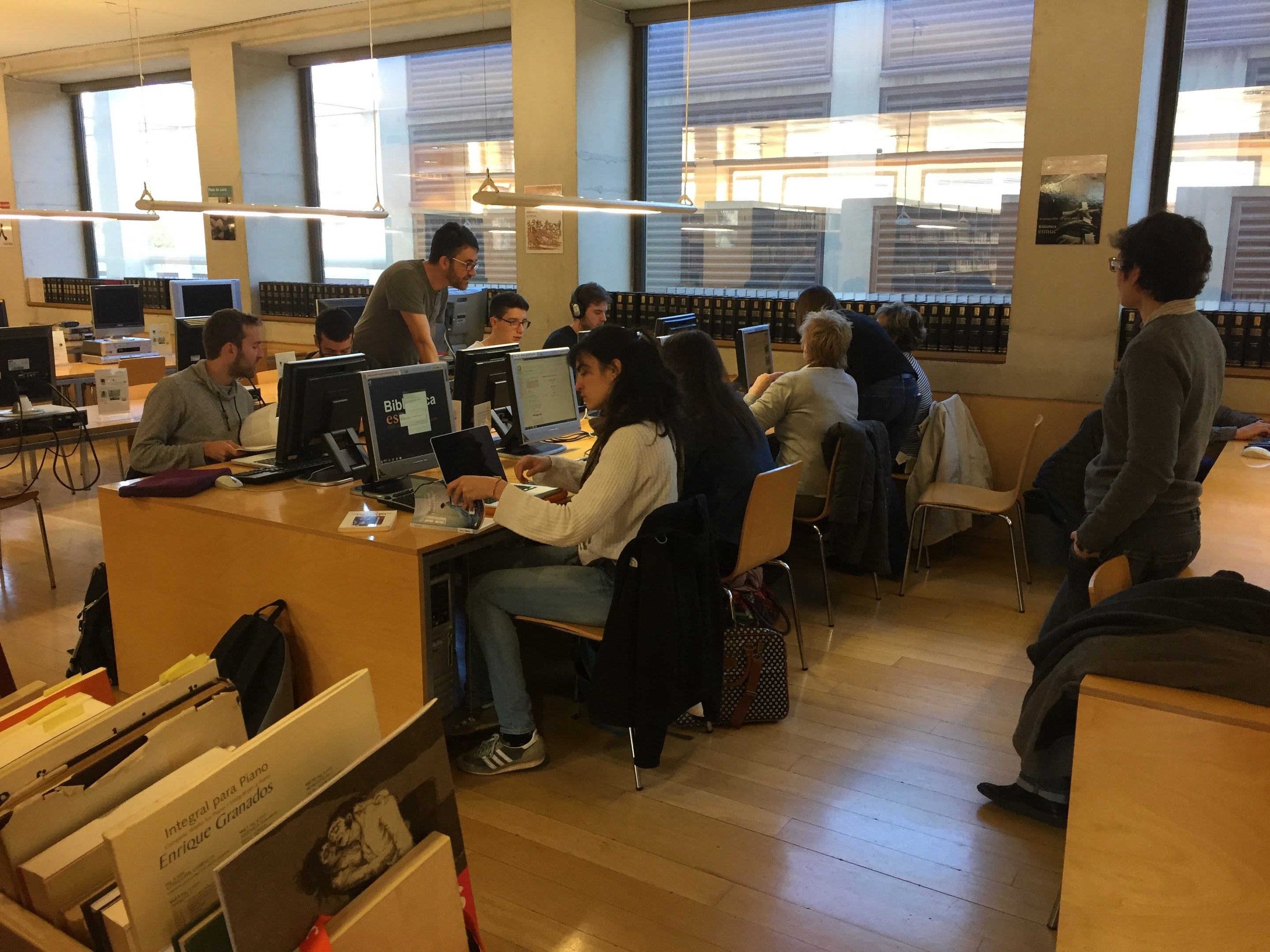 Eine Gruppe von Menschen, die an Computern auf Schreibtischen in einer Bibliothek mit Bücherregalen, Monitoren, Tastaturen, Mäusen, Taschen auf dem Boden, Deckenlampen und Glasfenstern im Hintergrund arbeiten.