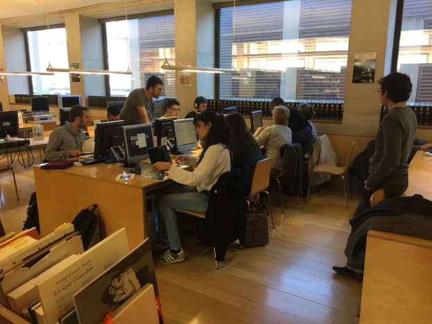Eine Gruppe von Menschen, die an Computern auf Schreibtischen in einer Bibliothek mit Bücherregalen, Monitoren, Tastaturen, Mäusen, Taschen auf dem Boden, Deckenlampen und Glasfenstern im Hintergrund arbeiten.