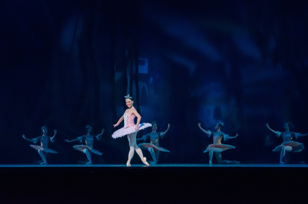 Eine Balletttänzerin in einem rosafarbenen Tutu und Spitzenschuhen tanzt anmutig auf der Bühne, umgeben von anderen gleich gekleideten Tänzerinnen, mit einem dunklen Hintergrund, der eine dramatische Atmosphäre schafft.