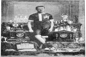 Schwarzes und weißes Foto eines Mannes in einem Fußballtrikot vor einem Tisch mit Pokalen und Trophäen, beschriftet mit "1909-1910 WM-Sieger."