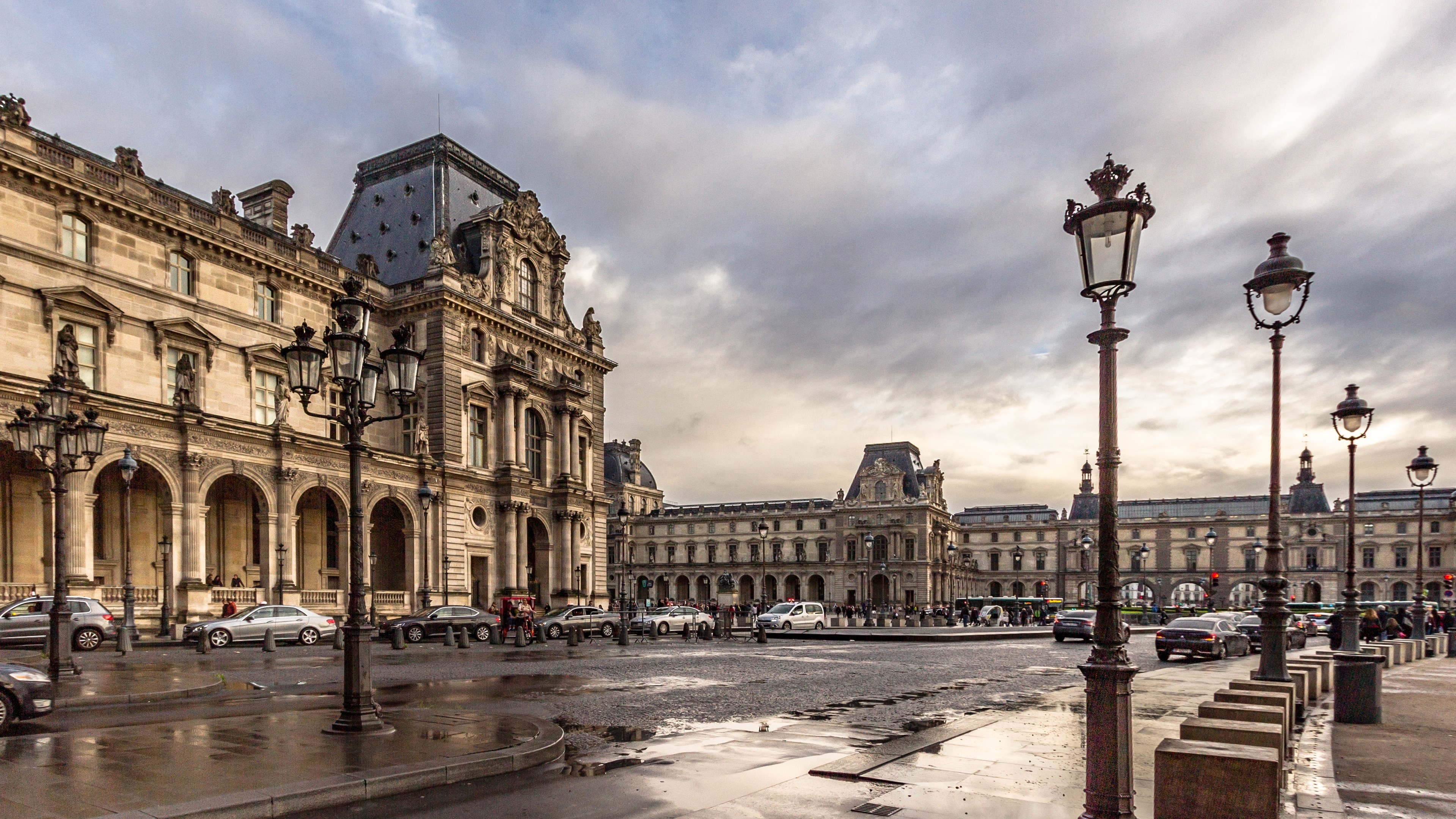 Außenansicht des Louvre-Museums in Paris, das seine charakteristische Architektur, Straßenlaternen, Lichter, Fahrzeuge, Passanten und einen bewölkten Himmel zeigt.