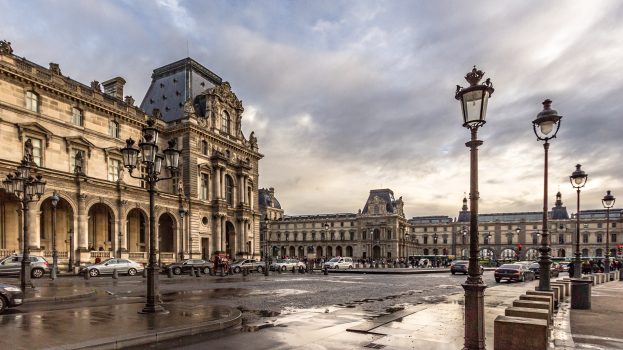 Außenansicht des Louvre-Museums in Paris, das seine charakteristische Architektur, Straßenlaternen, Lichter, Fahrzeuge, Passanten und einen bewölkten Himmel zeigt.