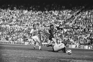 Schwarzes Foto von Fussballspielern im Einsatz auf einem Feld mit Zuschauern im Hintergrund und einem Banner "Bundesliga 1978-79 Ajax 1-1 Hannover 96"