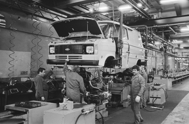 Eine Gruppe von Männern, die an einem Van in einer Fabrik mit Maschinen, Tischen und anderen Gegenständen arbeiten, mit Wänden, die mit Rohren und Lichtern versehen sind, in Schwarz-Weiß dargestellt.