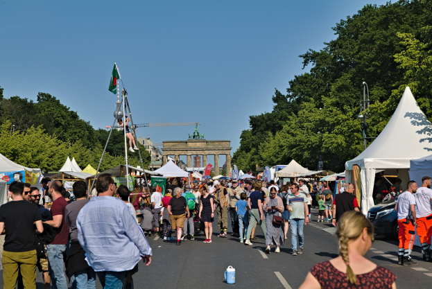 Eine Menschenmenge, die eine Straße mit Zelten, Fahrzeugen und Bäumen entlanggeht, mit einem Bogen und einem klaren blauen Himmel im Hintergrund und Polen mit Fahnen auf der linken Seite, wahrscheinlich das Oktoberfest in München, Deutschland.