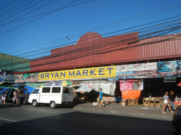 Ein belebter Marktplatz mit Fahrzeugen, Passanten, Marktständen, Bannern, Schirmen, hölzernen Gegenständen und einem 'Bryan Market'-Gebäude im Hintergrund unter einem Himmel mit Oberleitungen.