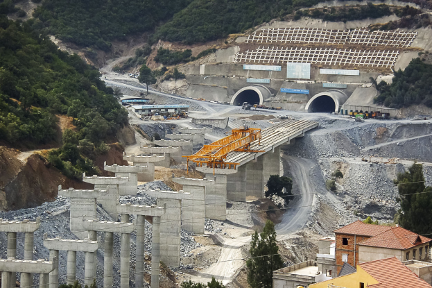 Großer Tunnel auf einer Baustelle umgeben von Bäumen, Gebäuden, Fahrzeugen, Säulen, Felsen und einer Straße unten.