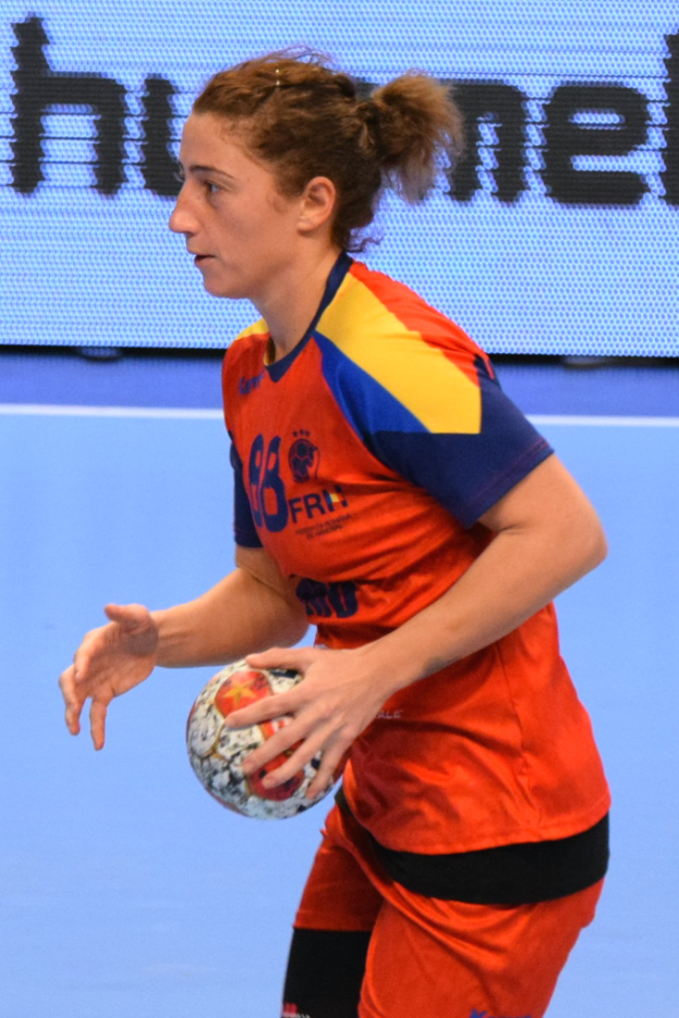 Eine Frau in einem orangen und blauen Outfit hält einen Fußball vor einem Bildschirm mit Text.