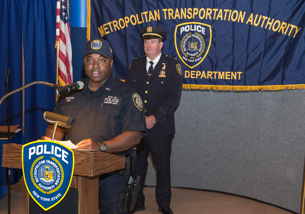 Zwei Polizeibeamte stehen vor einem Rednerpult, einer hölt Papiere und spricht in ein Mikrofon, mit einer Fahne, einem Tisch und einer Tafel mit Symbolen und Text in der Nähe.