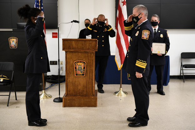 Eine Gruppe uniformierter Personen in Masken steht vor einem Podium mit einem Mikrofon, umgeben von Fahnen, Stühlen und Tischen, wobei eine Person ein Zertifikat hält, während es scheint, dass eine Vereidung eines neuen Feuerwehrchefs stattfindet.