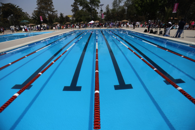 Großes Schwimmbad mit Bahnentrennung, umgeben von Menschen mit Taschen und Gegenständen, mit Zelten, Schildern, Stangen, einem Schild und einer bewölkten Himmel im Hintergrund.