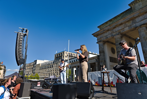 Gruppe von Menschen, die auf einer Bühne vor dem Brandenburger Tor spielen, begleitet von Equipment und städtischer Kulisse.
