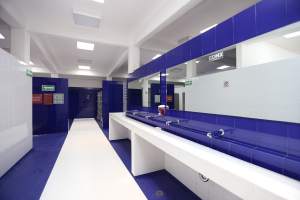 Öffentliche Toilette mit blauen Fliesenwänden, weißen Tresen, Waschbecken mit Wasserhähnen, einer Plattform mit einer Flasche und Gegenständen, Texttafeln an den Wänden, Deckenleuchten und Säulen.