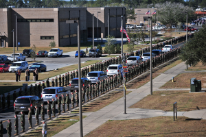Eine große Gruppe von Menschen in militärischer Uniform mit Gewehren vor einem Gebäude während einer Parade, mit Fahrzeugen, Laternenmasten, Fahnen, Bäumen und Gras im Vordergrund und Gebäuden unter einem klaren blauen Himmel im Hintergrund.