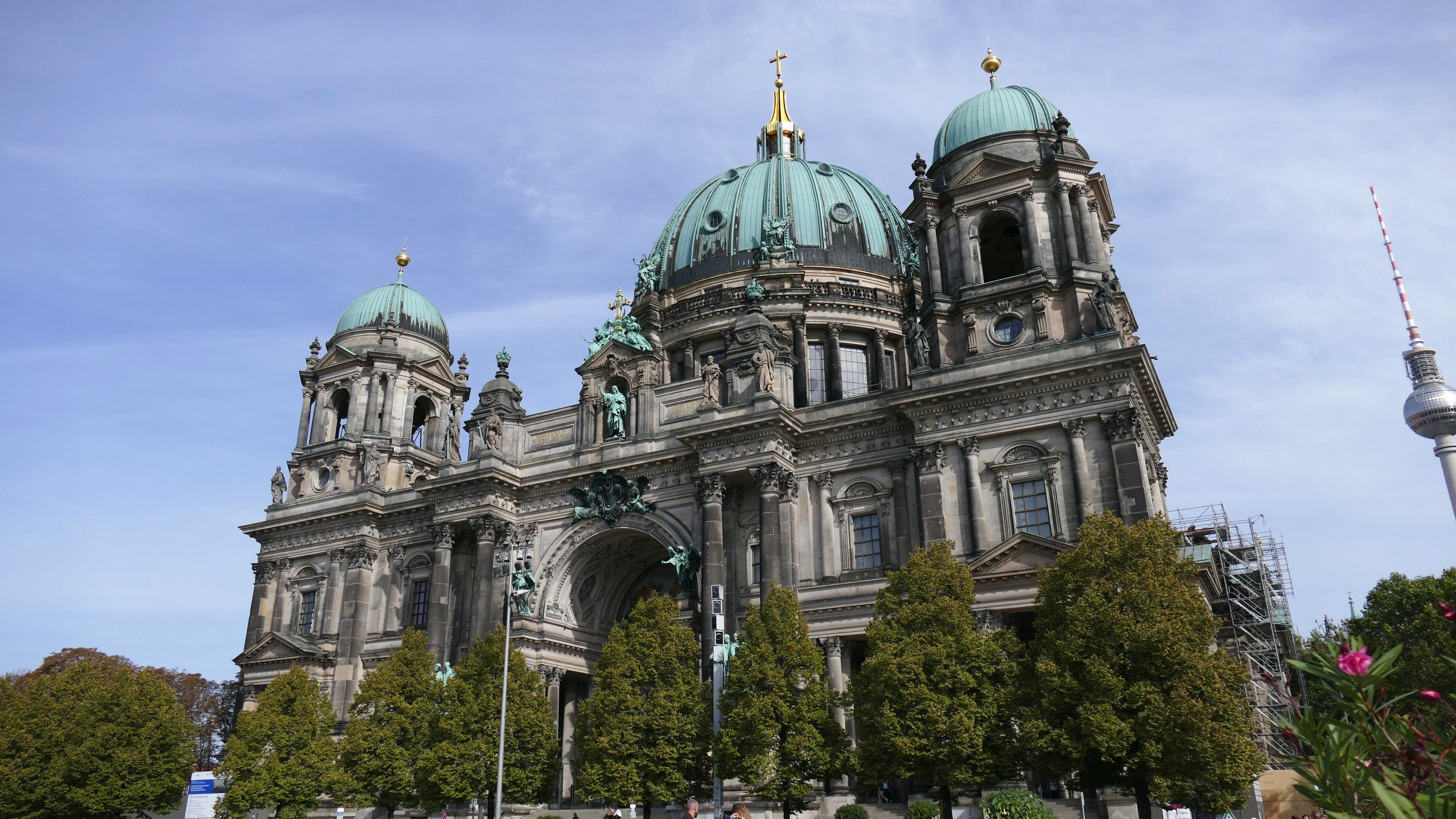Großes, prunkvolles Berliner Dom-Gebäude in Berlin, Deutschland, mit architektonischen Details wie Säulen, Bögen und Statuen, vor einem bewölkten Himmel, mit Passanten, Bäumen und einem Turm im Vordergrund.