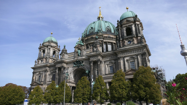 Großes, prunkvolles Berliner Dom-Gebäude in Berlin, Deutschland, mit architektonischen Details wie Säulen, Bögen und Statuen, vor einem bewölkten Himmel, mit Passanten, Bäumen und einem Turm im Vordergrund.